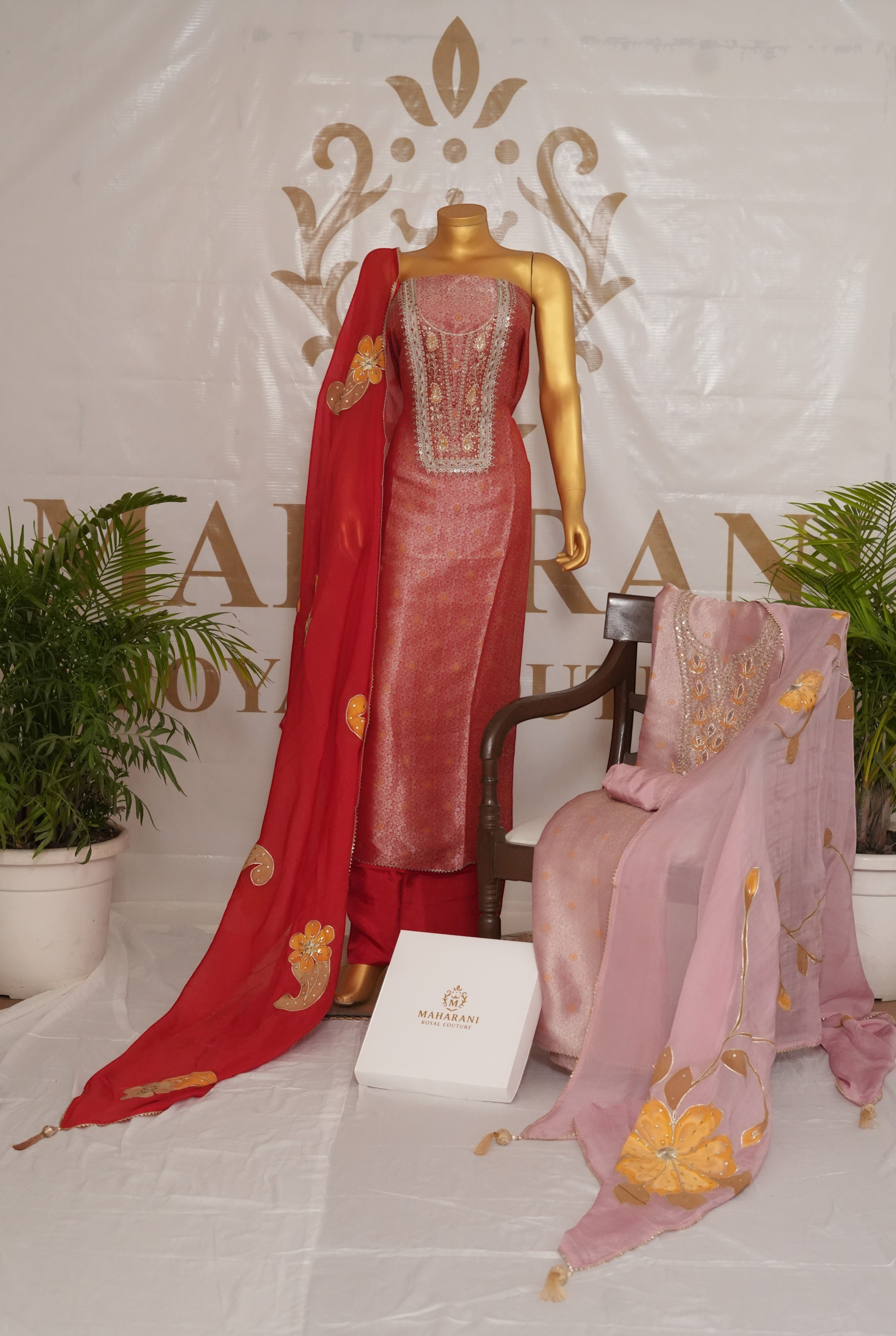 Maharani Royal Couture red Banarasi silk suit with hand embroidery, sequins, pearls, and red organza dupatta with painted motifs. Another suit in pink is draped over a chair.